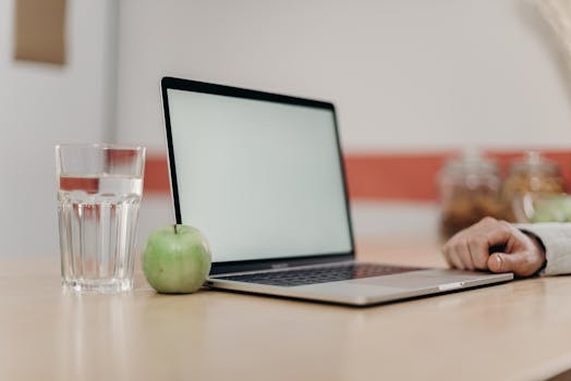 A clean and minimal home workspace with a laptop, green apple, and a glass of water on a desk.