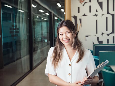 Asian woman smiling with a clipboard in hand, standing in a modern office environment.