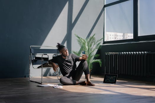 Man sitting on the floor, checking smartphone, with laptop showing stock market trends nearby.