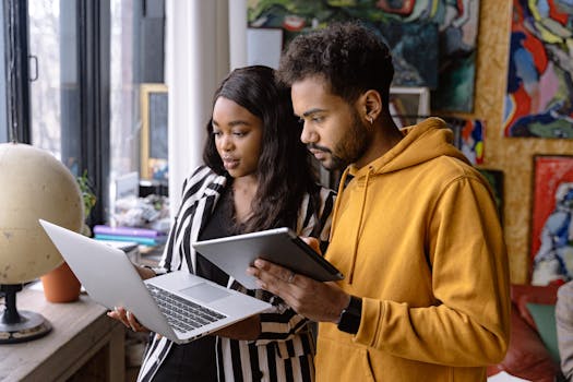 Two young adults collaborating using laptop and tablet indoors, showcasing teamwork and technology.