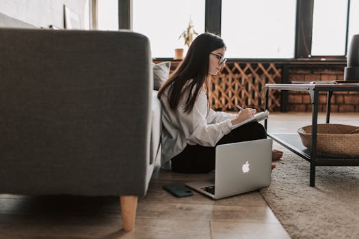 Woman sitting on floor with laptop and notebook, enjoying remote work in a comfortable home environment.