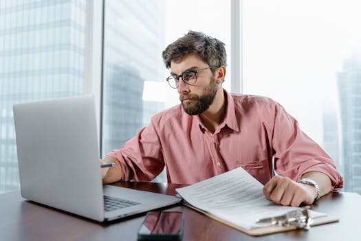 A businessman in an office reviews documents and data on a laptop, concentrating on financial analysis.