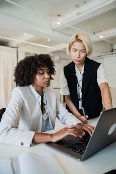 A mentor guiding an intern at a laptop in a bright, modern office setting. Professional teamwork emphasized.