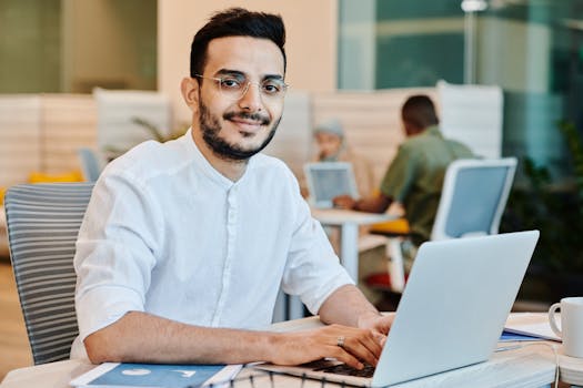 Asian man in eyeglasses using a laptop in a bright, modern workspace, focusing on tasks.