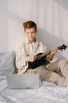 Redheaded preteen boy in beige outfit playing a ukulele while learning from a laptop at home.